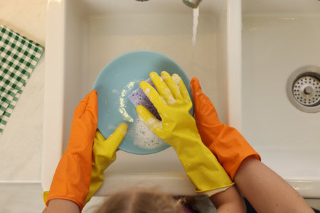 Mother and daughter in protective gloves washing plate above sink, top viewの写真素材