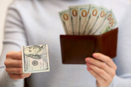 Woman giving 100 dollar bill and holding brown leather wallet with banknotes, closeup. money...