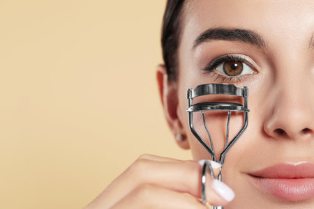 Beautiful young woman using eyelash curler on beige background, closeup. Space for textの写真素材