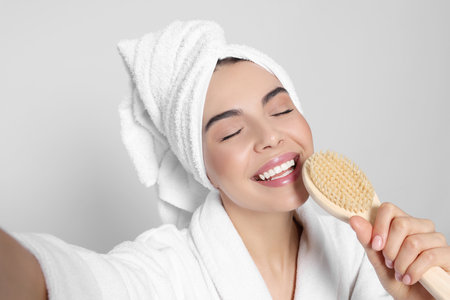 Woman in bathrobe with towel taking selfie while singing into brush on light gray background. spa treatmentの写真素材