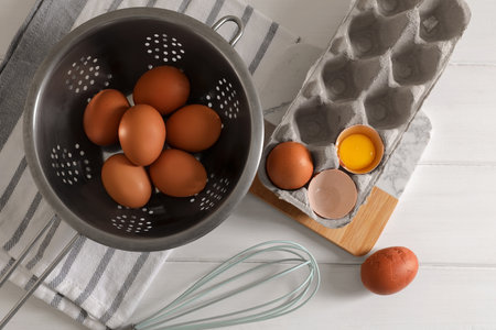 Fresh raw chicken eggs with colander, napkin and whisk on white wooden table, flat layの写真素材