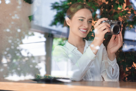 Young woman with camera taking photo at cafe, view through window. creative hobby