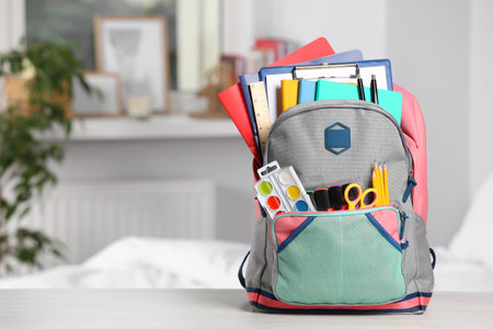 Backpack with different school stationery on white table indoors, space for text