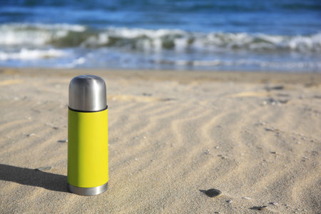 Yellow flask with hot drink on sand near sea, space for textの写真素材