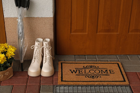 Door mat with word Welcome, boots, umbrella and beautiful flowers near entranceの写真素材