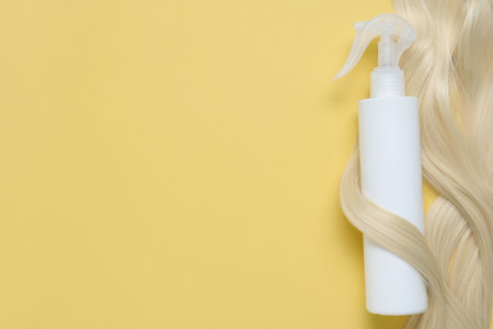 Spray bottle with thermal protection wrapped in lock of blonde hair on yellow background, top view. Space for textの写真素材