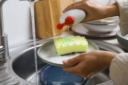 Woman pouring dishwashing detergent onto plate with sponge near kitchen sink, closeupの写真素材