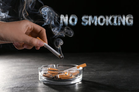 No Smoking, phrase of smoke. Woman holding smoldering cigarette over glass ashtray at gray table against black background, closeupの写真素材