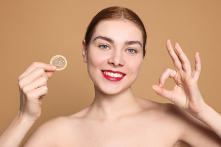 Woman with condom showing ok gesture on beige background. safe sexの写真素材