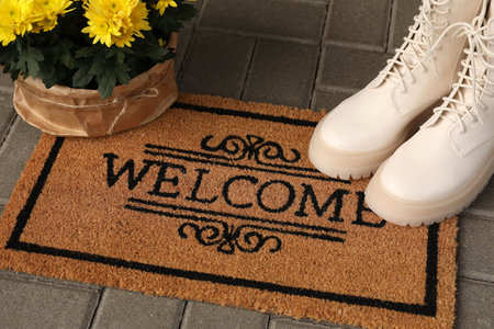 Door mat with word Welcome, stylish boots and beautiful flowers on floorの写真素材