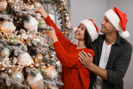 Happy young couple in Santa hats decorating Christmas tree together at homeの写真素材