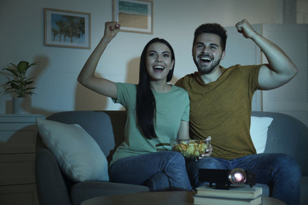 Emotional young couple watching TV using video projector at homeの写真素材