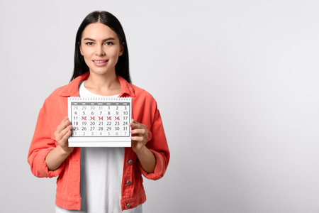Young woman holding calendar with marked menstrual cycle days on light background. Space for textの写真素材
