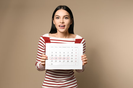 Young woman holding calendar with marked menstrual cycle days on beige backgroundの写真素材