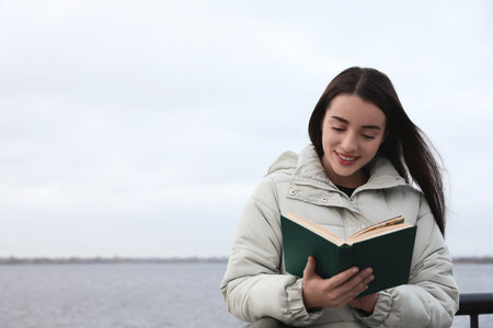 Woman reading book near river on cloudy dayの写真素材