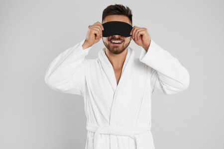 Young man in bathrobe and eye sleeping mask on light gray backgroundの写真素材