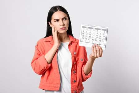 Pensive young woman holding calendar with marked menstrual cycle days on light backgroundの写真素材