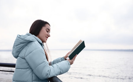 Woman reading book near river on cloudy dayの写真素材