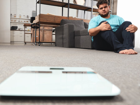 Depressed overweight man looking at scales in living roomの写真素材
