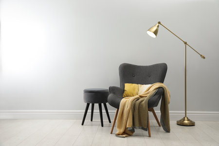 Stylish armchair with plaid, floor lamp and ottoman near white wall, space for text. interior designの写真素材