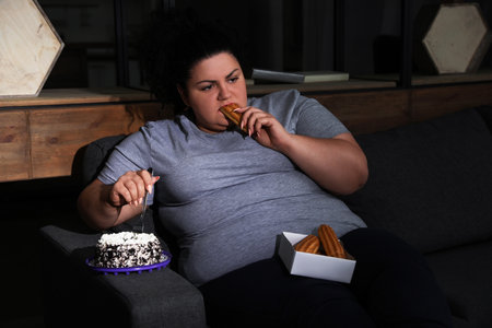 Depressed overweight woman eating sweets in living room at nightの写真素材