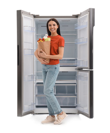 Young woman with bag of groceries near empty refrigerator on white backgroundの写真素材
