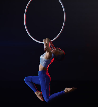 Young woman performing acrobatic element on aerial ring against dark backgroundの写真素材