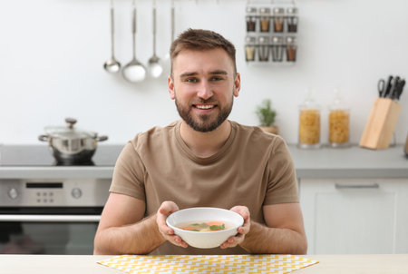Young man holding bowl of tasty soup at table in kitchenの写真素材