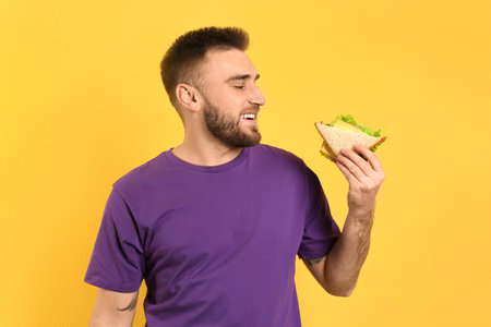 Young man eating tasty sandwich on yellow backgroundの写真素材