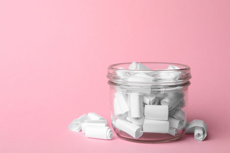 Glass jar full of rolled paper sheets on pink background, space for text