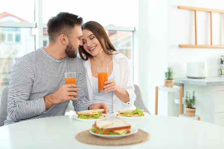 Happy couple having breakfast with sandwiches at table in kitchenの写真素材