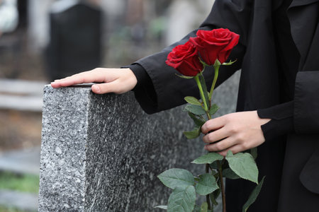 Woman with red roses near gray granite tombstone outdoors, closeup. Funeral ceremonyの写真素材