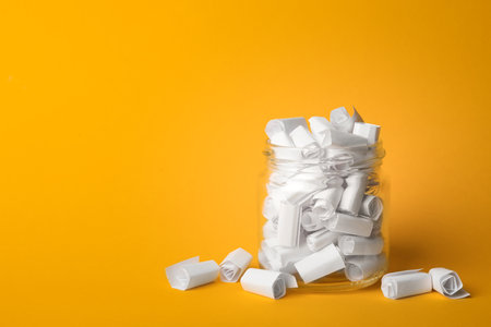 Glass jar full of rolled paper sheets on yellow background, space for text