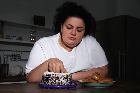 Depressed overweight woman eating cake in kitchen at nightの写真素材