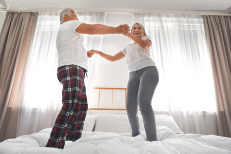 Happy mature couple dancing together on bed at home, low angle viewの写真素材