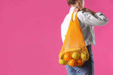 Woman holding net bag with fruits on pink background, closeup. Space for textの写真素材