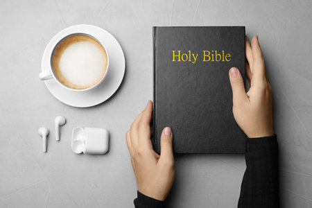 Woman with Bible, cup of coffee and earphones at light gray table, top view. religious audiobook