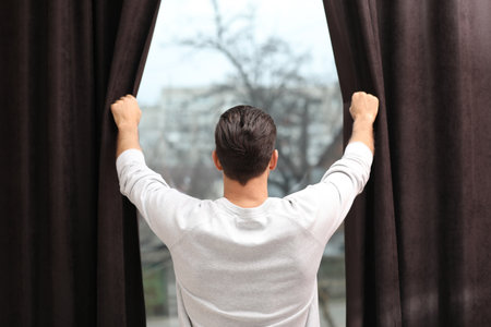 Man opening window curtains at home, back viewの写真素材