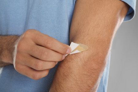 Man putting sticking plaster onto arm against light gray background, closeup
