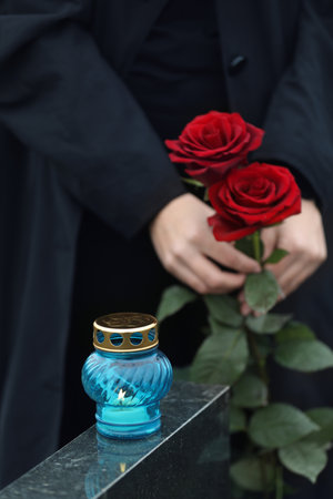 Woman holding red roses near black granite tombstone with candle outdoors, closeup. Funeral ceremonyの写真素材