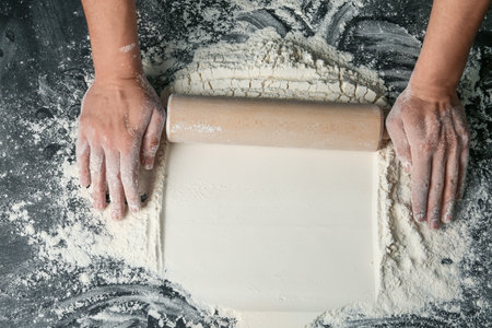 Baker rolling white flour with pin on dark table, closeup. Space for text