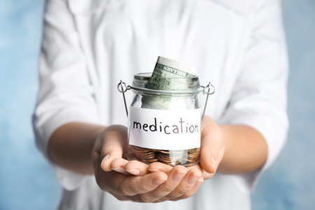 Woman holding glass jar with money and tag MEDICATION on light blue background, closeupの写真素材
