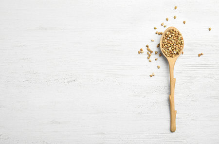 Uncooked green buckwheat grains in spoon on white wooden table, top view. Space for textの写真素材