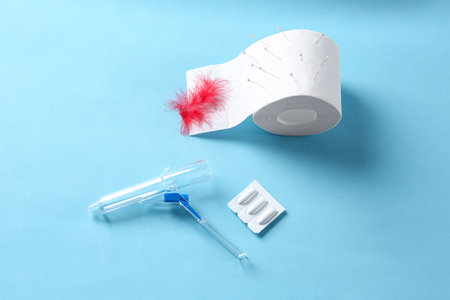 Anoscope, suppositories and toilet paper with red feather on light blue background, flat lay....