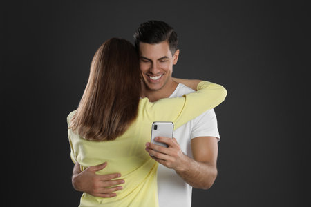 Man interested in smartphone while hugging his girlfriend on black background. relationship problemsの写真素材