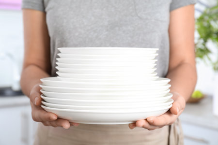Woman holding stack of clean white plates in kitchen, closeup