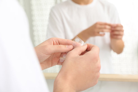 Teen guy using acne healing patch near mirror indoors, focus on handsの写真素材