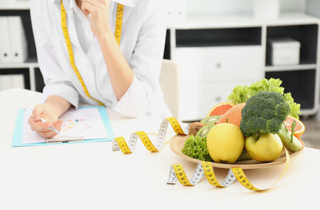 Nutritionist at desk with fruits, vegetables and measuring tape in clinic, closeupの写真素材