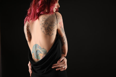 Beautiful woman with tattoos on body against black background, back view. Space for textの写真素材
