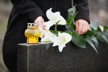 Woman holding white lilies near black granite tombstone with candle outdoors, closeup. Funeral ceremonyの写真素材
